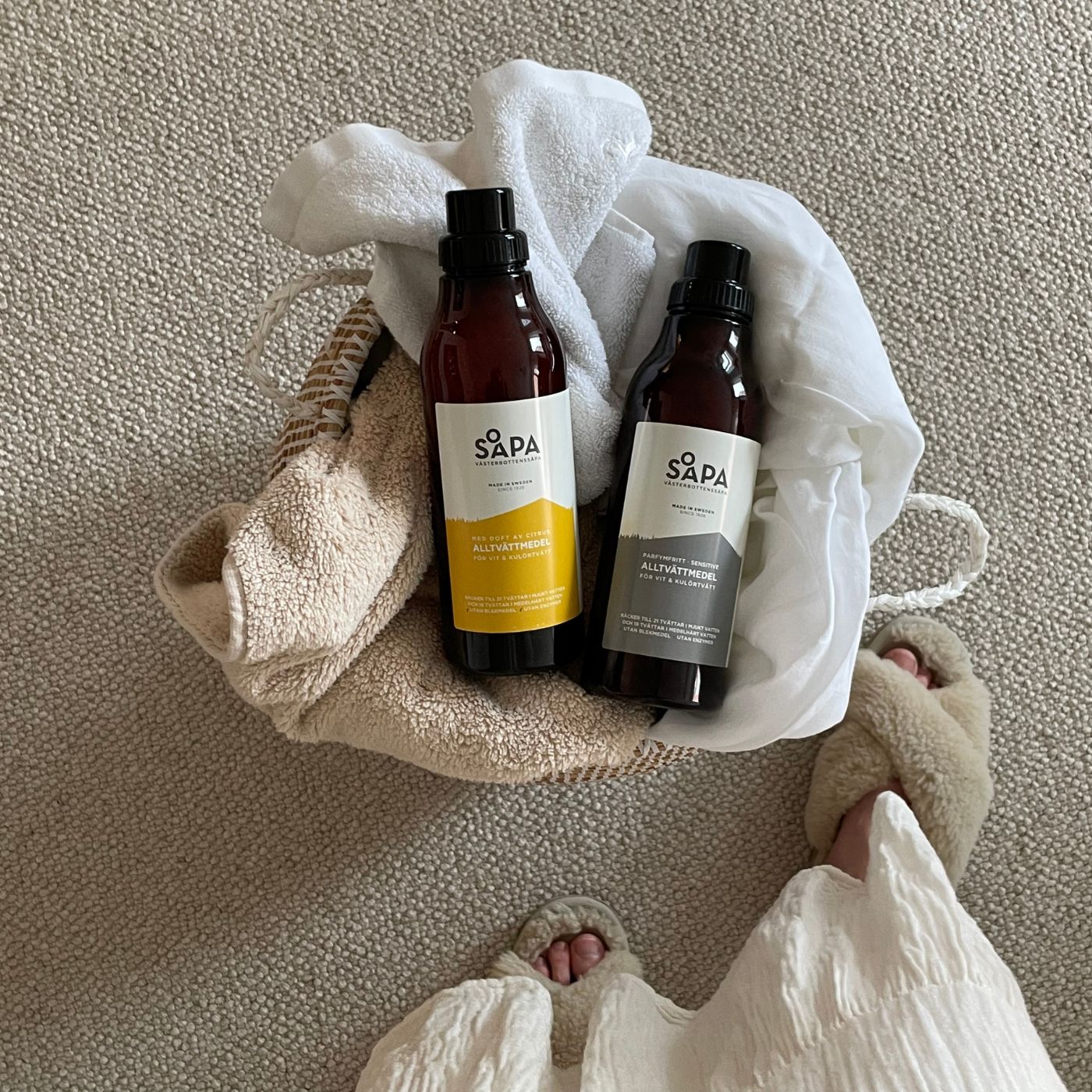 Två flaskor av Västerbottenssåpas tvättmedel - en med fräsch citrondoft och en för känslig hud utan parfym - i en korg med handdukar